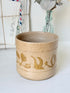 A handmade ceramic planter in a sandy beige stoneware finish with a glossy gold leaf pattern, set against a softly blurred background of vintage mail and a glass bottle vase.
