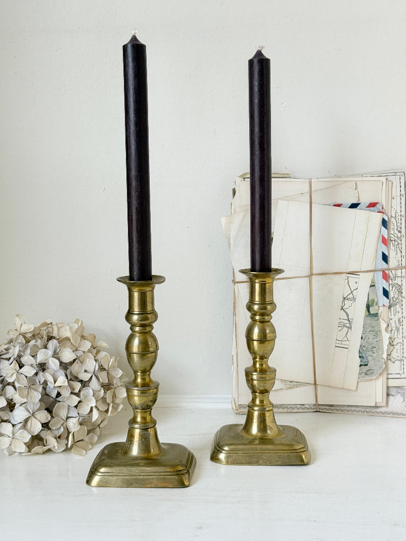 Pair of turned brass candlesticks with square stepped bases and black taper candles on a white surface, styled with dried flowers