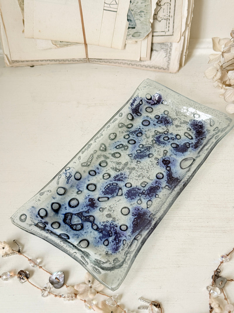 A rectangular glass serving tray with a textured, bubbly pattern in shades of blue, purple, and gray, styled on a light wooden surface with vintage papers and jewelry in the background.