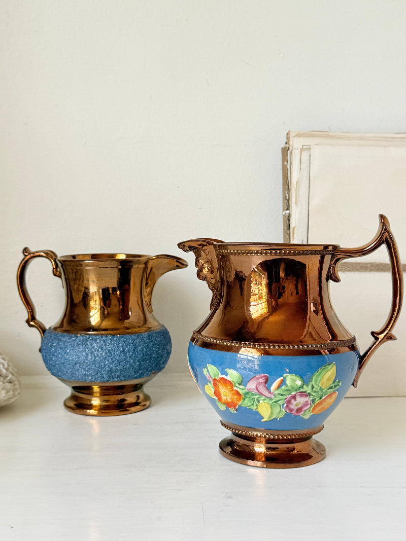 Two vintage-style metal pitchers on a white surface against a white wall. The pitcher on the left is copper-toned with a textured blue lower section.