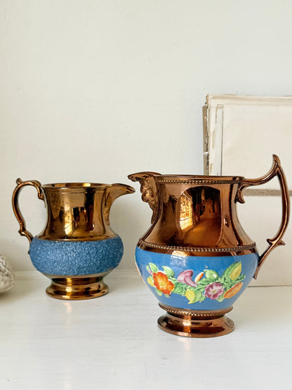 Two vintage-style metal pitchers on a white surface against a white wall. The pitcher on the left is copper-toned with a textured blue lower section.