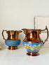 Two vintage-style metal pitchers on a white surface against a white wall. The pitcher on the left is copper-toned with a textured blue lower section.