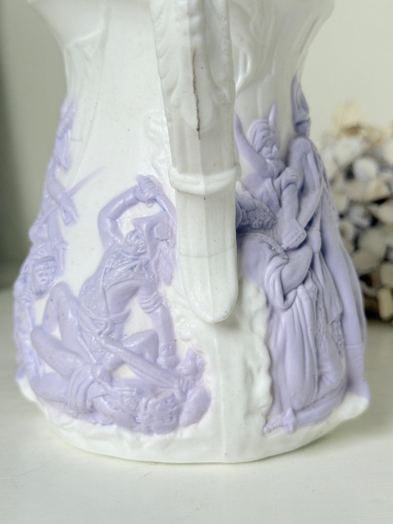 A close-up of a white ceramic pitcher decorated with pale lavender relief figures and scenes, showing the handle and part of the body against a softly blurred light background.