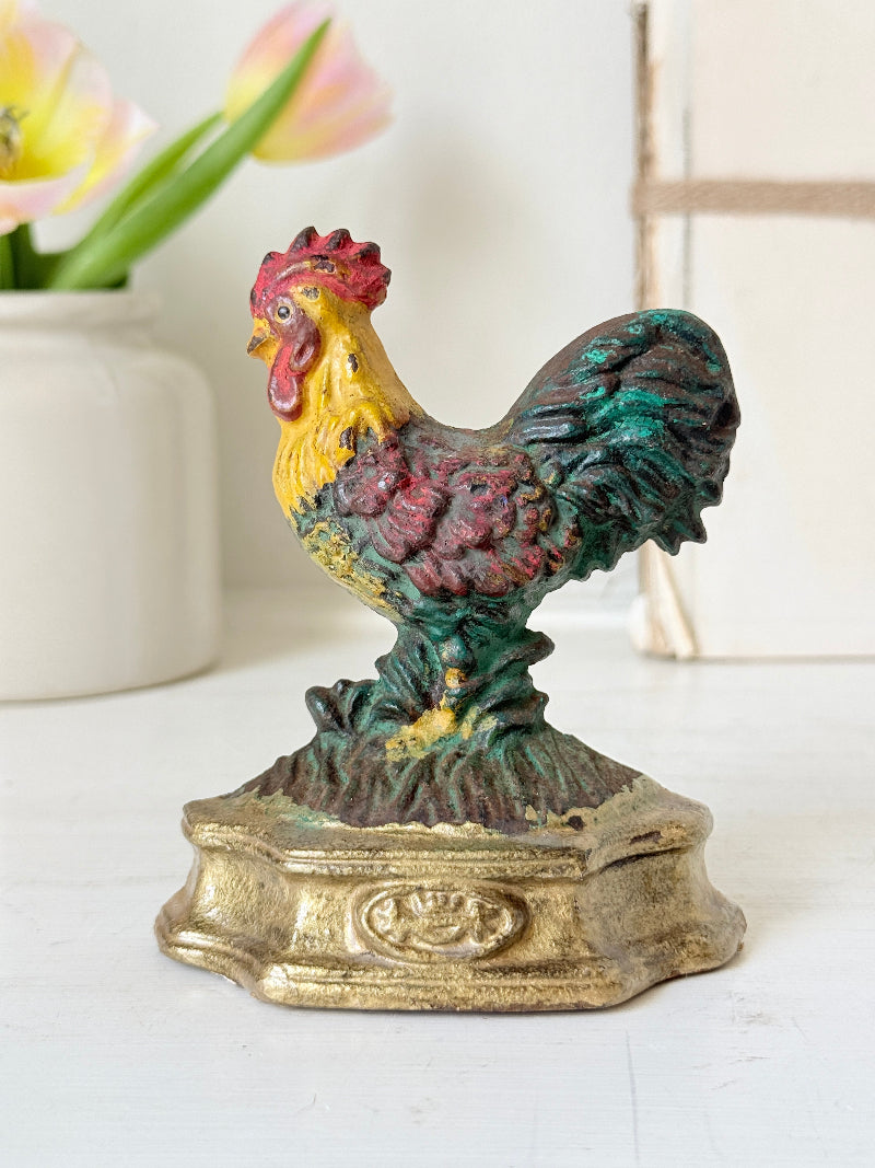 A vintage cast iron rooster bank or figurine on an ornate gold-colored base, set against a softly blurred background with a white vase of tulips.
