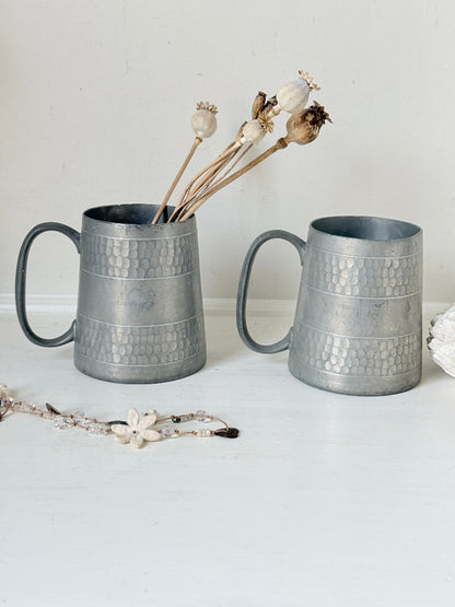 Pair of vintage pewter tankards with handles and textured detail, photographed on a white surface