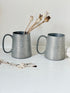 Pair of vintage pewter tankards with handles and textured detail, photographed on a white surface