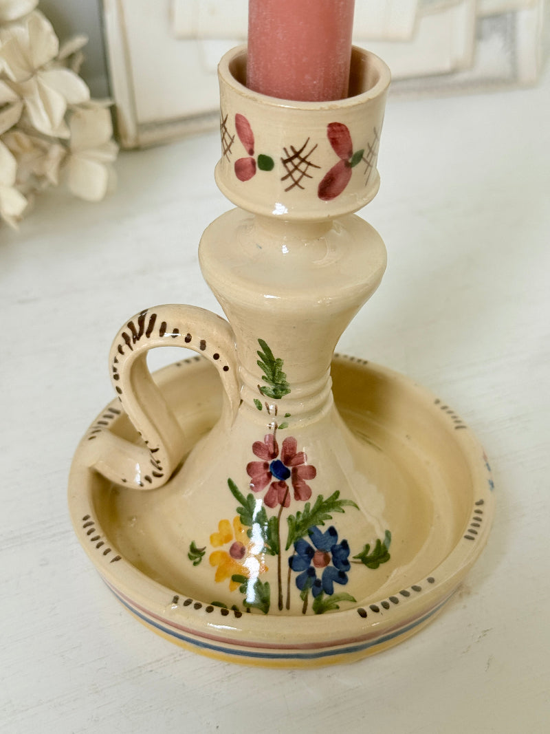 A vintage-style ceramic candlestick holder with a matching saucer base, featuring a cream-colored glaze and hand-painted floral motifs in pink, blue, yellow, and green. A pink candle is inserted into the top holder.
