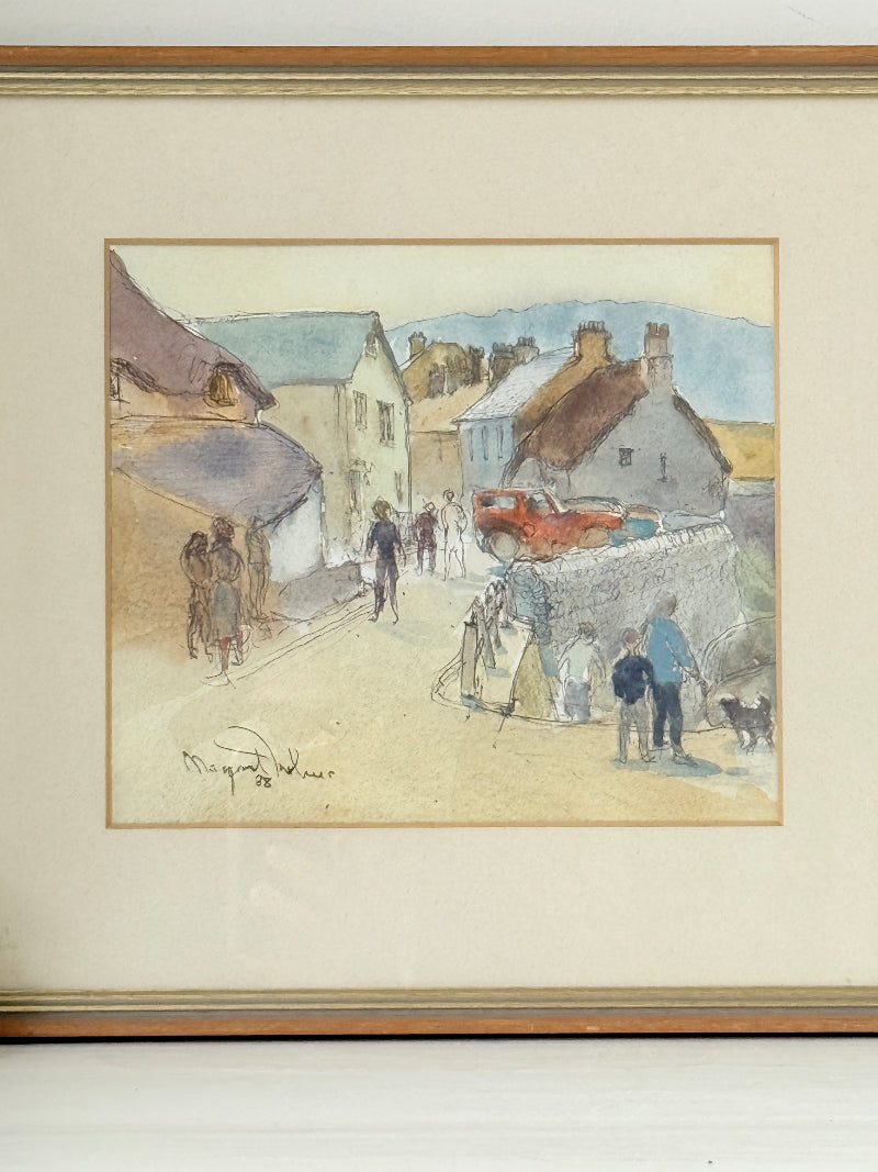 A framed watercolor painting depicting a village street scene with people, houses, and a red car, mounted with a cream mat and a gold-toned frame.