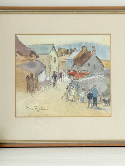 A framed watercolor painting depicting a village street scene with people, houses, and a red car, mounted with a cream mat and a gold-toned frame.