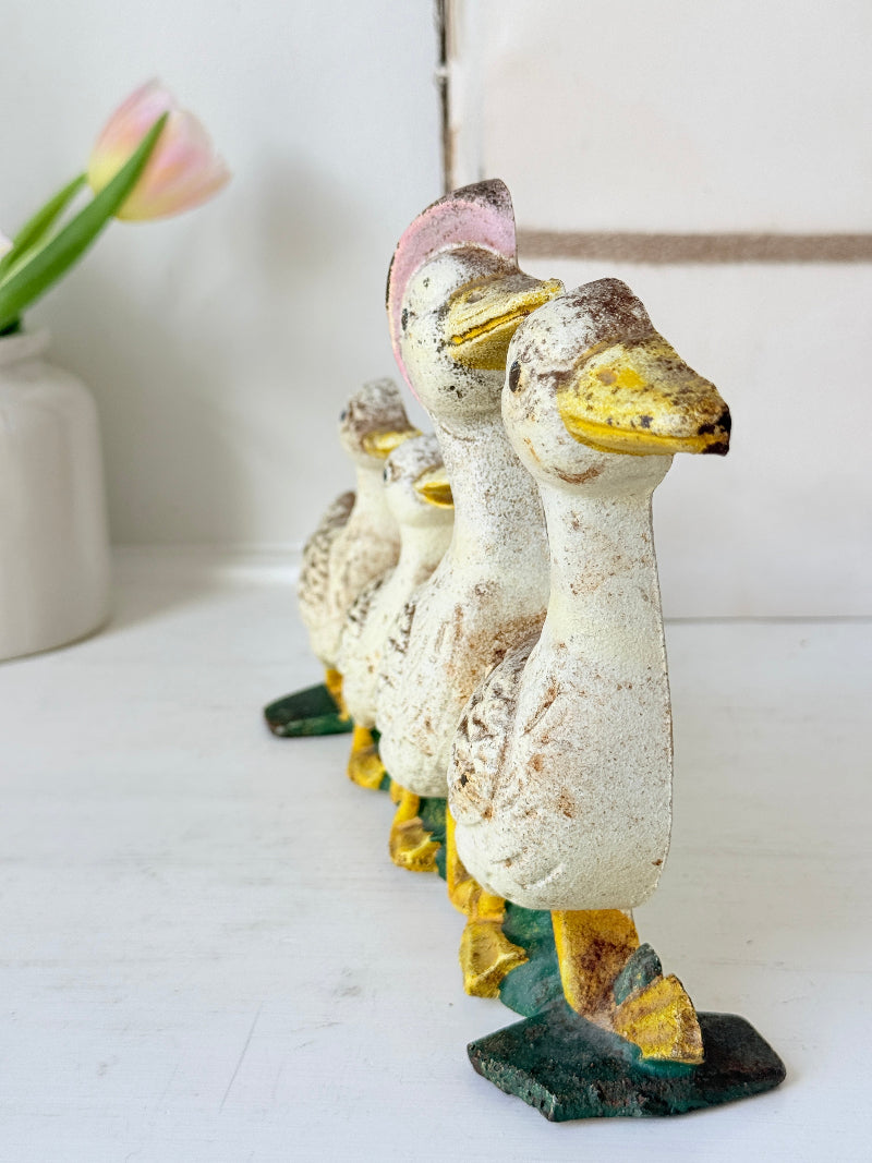 A set of five distressed cast-iron duck figurines lined up on a white surface, featuring a weathered cream finish with yellow, green, and pink accents.