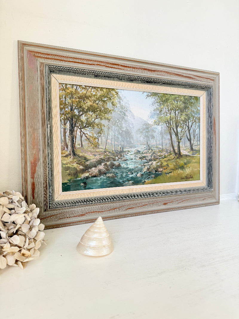 A framed landscape oil painting of a forest stream, displayed on a white surface next to a decorative shell and dried flowers. The frame is a distressed, rustic wood grain with a weathered silver and gold-toned inner border.