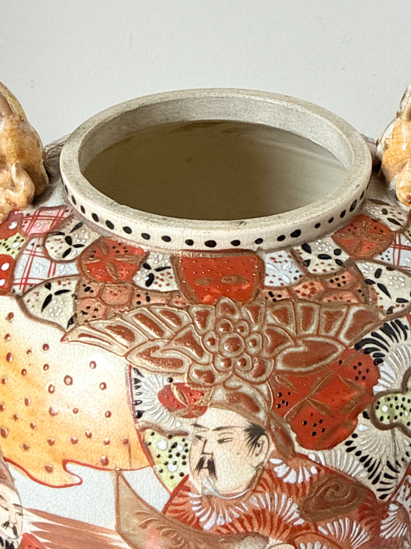 Close-up of a ceramic vase or tumbler with a wide rim, decorated with hand-painted Japanese-style figures and floral motifs in red, orange, beige, and black on a white glaze.