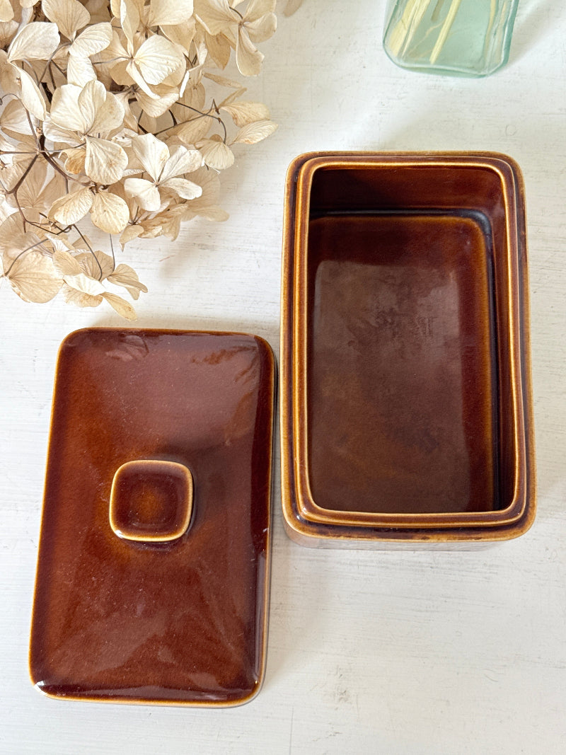 A glossy brown rectangular ceramic container with its matching lid placed beside it on a light surface. The container has a warm amber-brown glaze with a slightly lighter rim, and the lid features a small raised square knob.