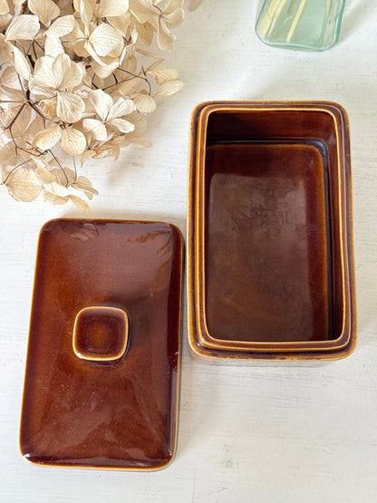 A glossy brown rectangular ceramic container with its matching lid placed beside it on a light surface. The container has a warm amber-brown glaze with a slightly lighter rim, and the lid features a small raised square knob.