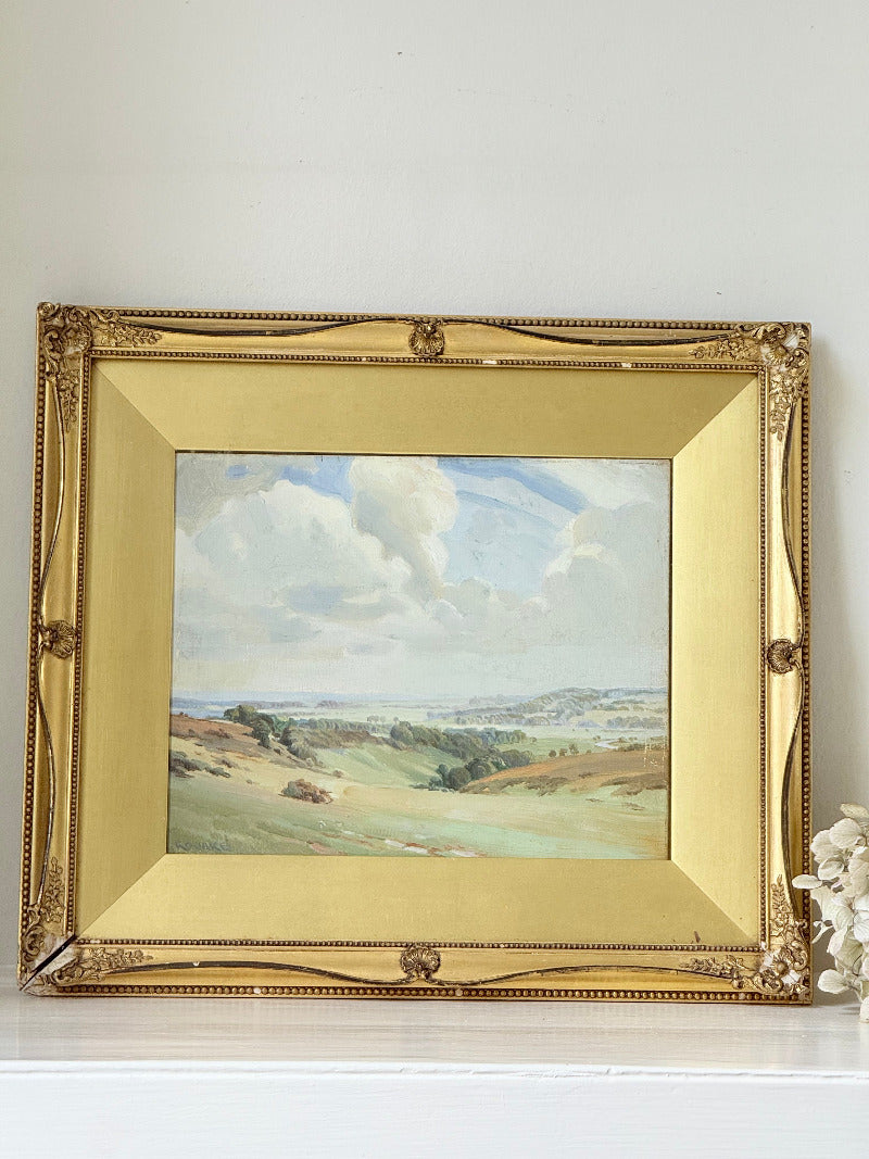 Framed oil painting of a rolling countryside landscape under a dramatic cloud-filled sky in an ornate gilt frame with wide mount.