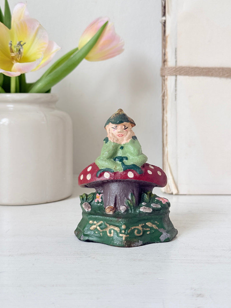 A small decorative gnome figurine sitting on a red-and-white mushroom cap, displayed on a white tabletop with a white vase of tulips and a rustic box in the background.