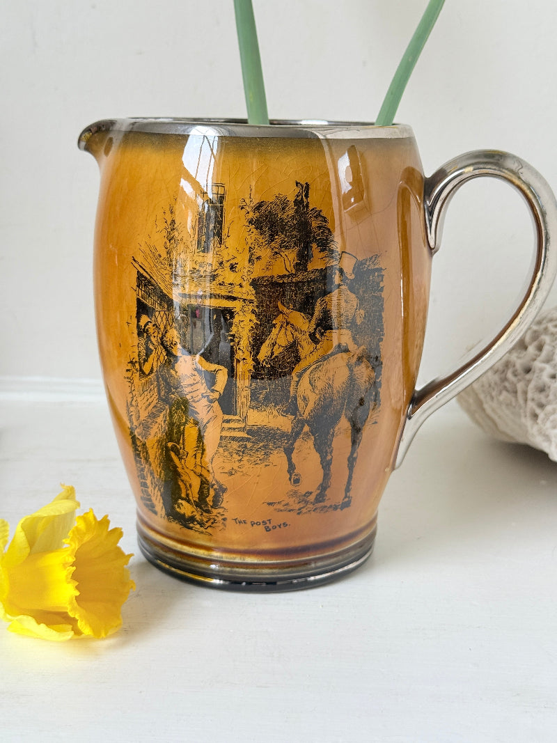Antique Ridgways stoneware jug photographed on a white surface in amber treacle glaze with black Coaching Days and Coaching Ways transfer scenes and silver tone handle