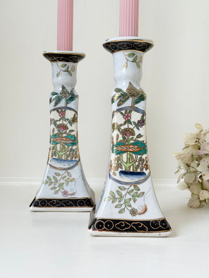 Pair of porcelain candle holders with hand-painted floral and vase motif in muted greens, pinks, and gold, styled with blush pink taper candles.