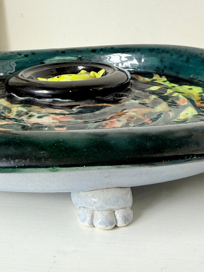 A close-up of a ceramic planter or decorative bowl with a glossy dark green glazed rim, a black central ring holding yellow-green leaves, and a white foot-shaped base supporting the piece.