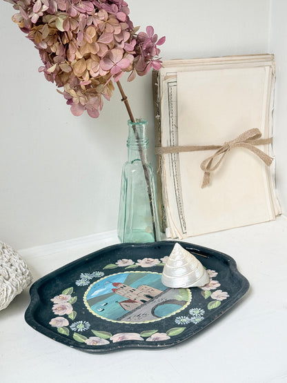 A decorative oval tray with scalloped edges, featuring a painted castle scene and pink floral border, styled on a white surface with a small white shell on top. A pale green glass bottle with dried pink flowers and a stack of cream books tied with twine are in the background.