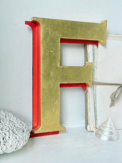 A large decorative letter F wall sign with a distressed gold top and red sides, styled on a white surface with a white crochet bag and a small clear shell.