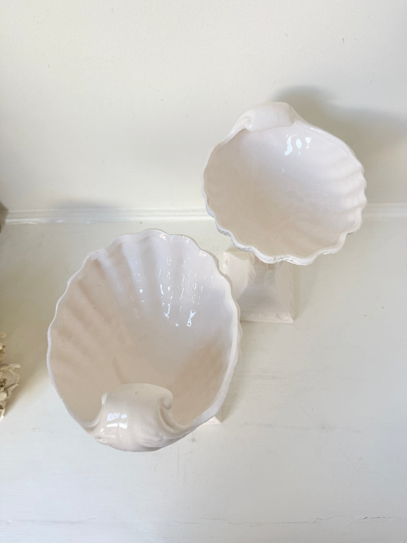 Two glossy white ceramic seashell-shaped bowls displayed on a white surface against a white wall.