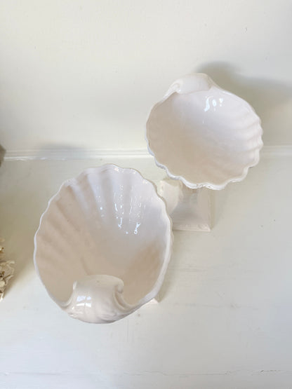 Two glossy white ceramic seashell-shaped bowls displayed on a white surface against a white wall.