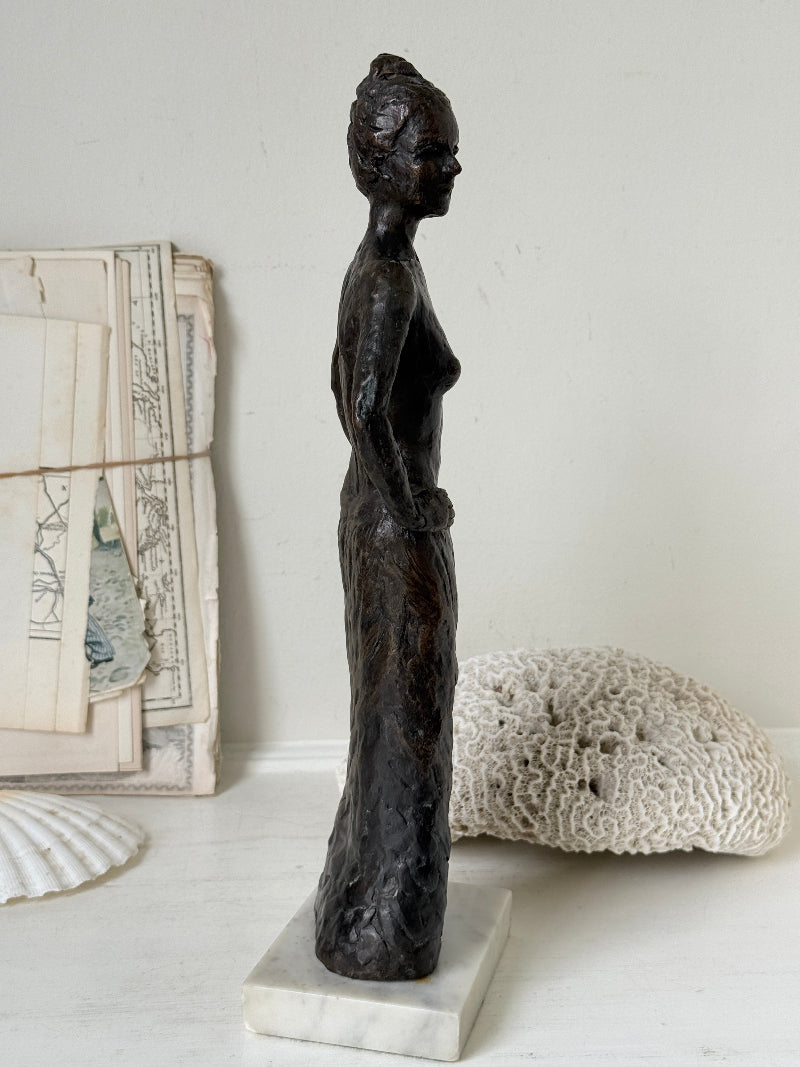 Solid bronze figure of a standing semi nude woman with upswept hair on a white marble base