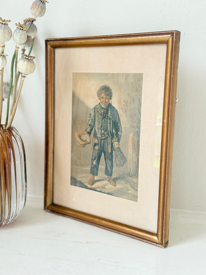 Vintage print of Little Beggar Boy in muted colours, framed under glass in simple wooden frame, on white surface.