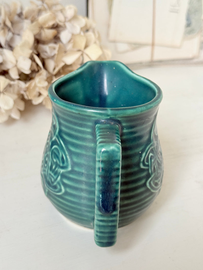 A small teal ceramic pitcher with a ribbed texture and embossed floral details, shown on a light surface with dried flowers in the background.