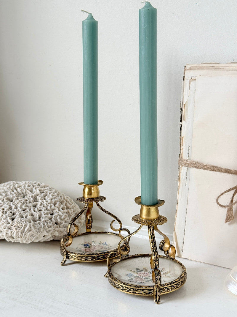 Two tall sage green taper candles in ornate gold-toned metal candlesticks with floral glass bases, styled on a white surface next to a lace doily and a vintage book.