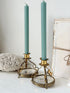 Two tall sage green taper candles in ornate gold-toned metal candlesticks with floral glass bases, styled on a white surface next to a lace doily and a vintage book.