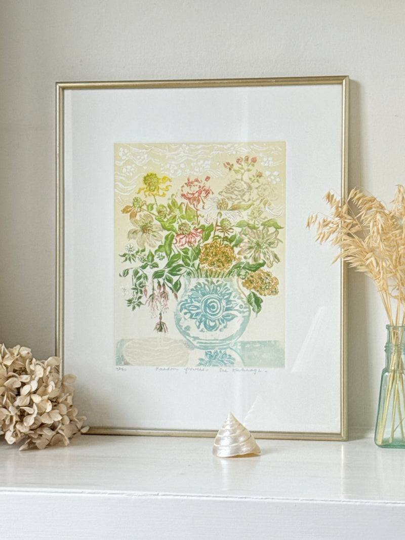 A framed botanical art print featuring a vintage-style floral bouquet in a blue patterned vase, displayed on a white mantel with dried flowers and a small shell.