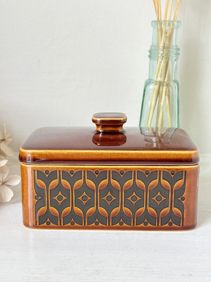 A rectangular ceramic butter dish with a lid, featuring a glossy brown and amber-orange glaze with a dark gray geometric pattern on the front, set against a light background with a glass reed diffuser bottle behind it.