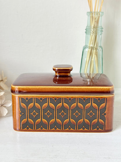 A rectangular ceramic butter dish with a lid, featuring a glossy brown and amber-orange glaze with a dark gray geometric pattern on the front, set against a light background with a glass reed diffuser bottle behind it.