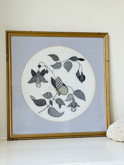 Framed circular hand embroidery of birds and fuchsia flowers in black and grey on white linen, set in gold frame, white surface background.