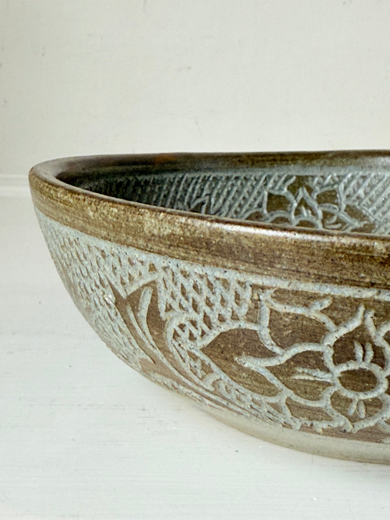 Studio pottery bowl with carved floral and geometric decoration in neutral glaze, photographed on a white surface with a pear inside.
