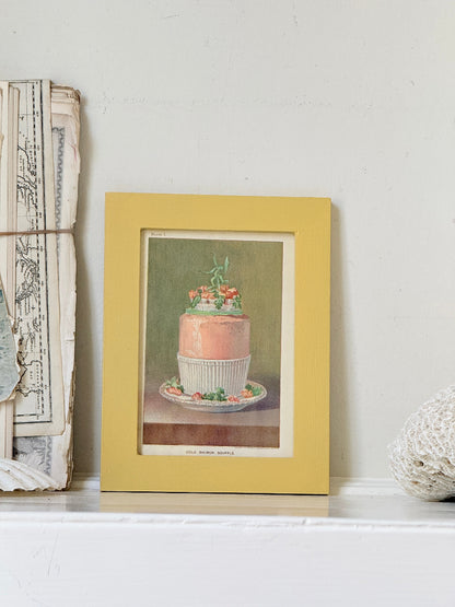 Framed Victorian cookery illustration of a celebration cake from 1896 in a mustard yellow frame displayed on a white surface.