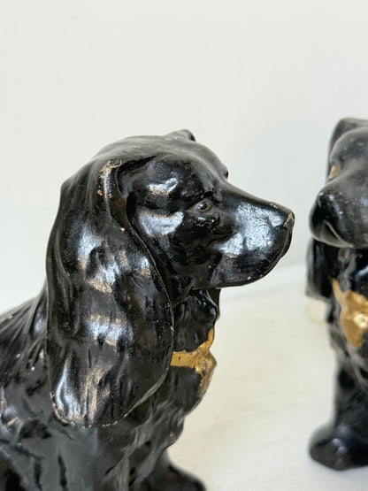 A close-up of a glossy black dog figurine with gold painted accents on its neck and chest, shown against a plain light background with part of another similar figurine visible on the right.