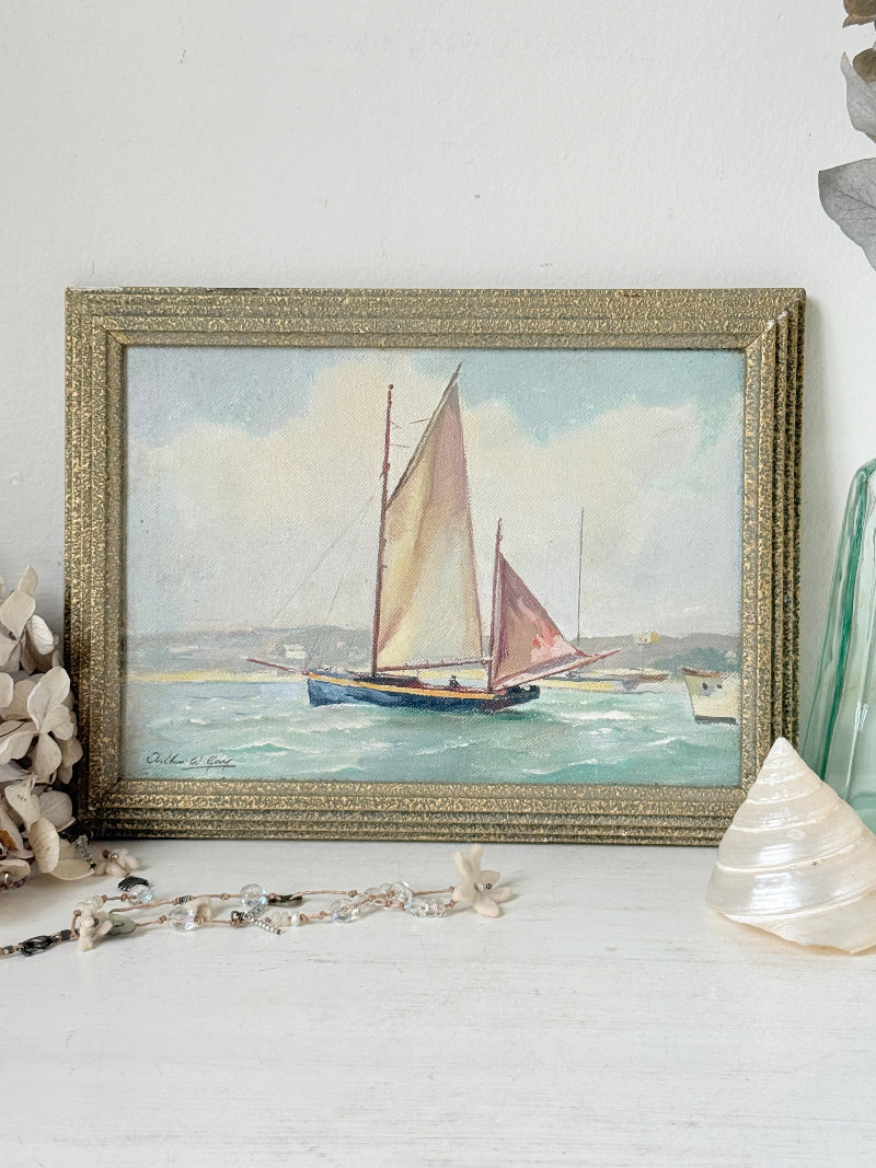 A framed vintage-style painting of a sailboat on the water, displayed on a white surface with decorative seashells and beads around it.