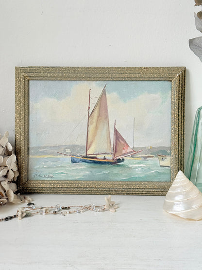 A framed vintage-style painting of a sailboat on the water, displayed on a white surface with decorative seashells and beads around it.