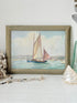 A framed vintage-style painting of a sailboat on the water, displayed on a white surface with decorative seashells and beads around it.