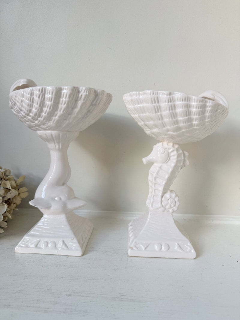 A pair of white ceramic nautical-themed candle holders, one shaped like a seashell on a pedestal and the other like a seahorse holding a shell, set against a neutral light gray background.