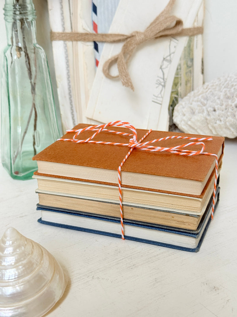 A stack of vintage hardcover books tied with red-and-white striped string, styled on a light tabletop with a glass bottle, seashells, and a white woven textile in the background.