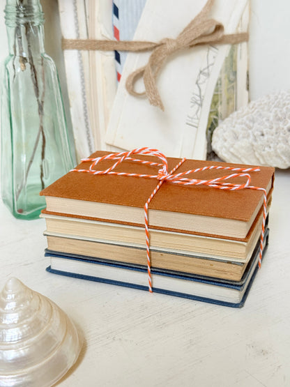 A stack of vintage hardcover books tied with red-and-white striped string, styled on a light tabletop with a glass bottle, seashells, and a white woven textile in the background.