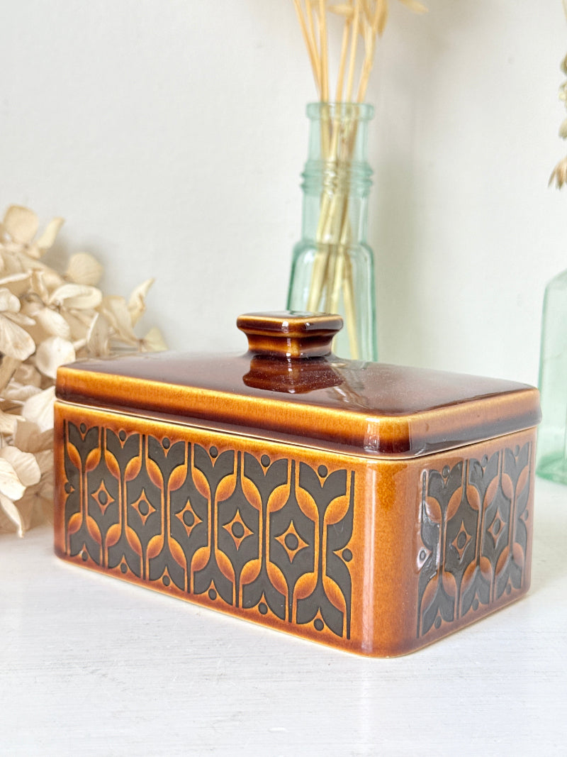 A rectangular ceramic butter dish with a lid, featuring a glossy amber-brown and black geometric pattern, styled on a white surface with dried flowers and glass bottles in the background.