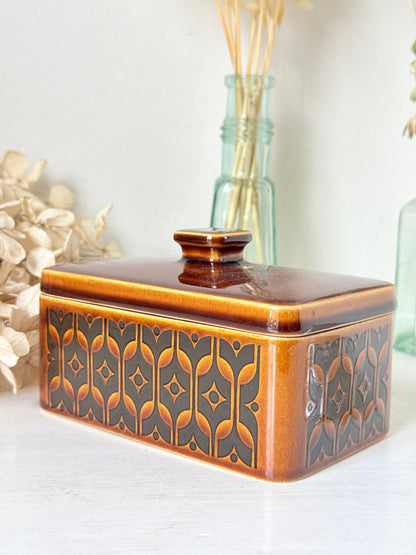 A rectangular ceramic butter dish with a lid, featuring a glossy amber-brown and black geometric pattern, styled on a white surface with dried flowers and glass bottles in the background.