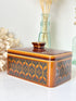 A rectangular ceramic butter dish with a lid, featuring a glossy amber-brown and black geometric pattern, styled on a white surface with dried flowers and glass bottles in the background.