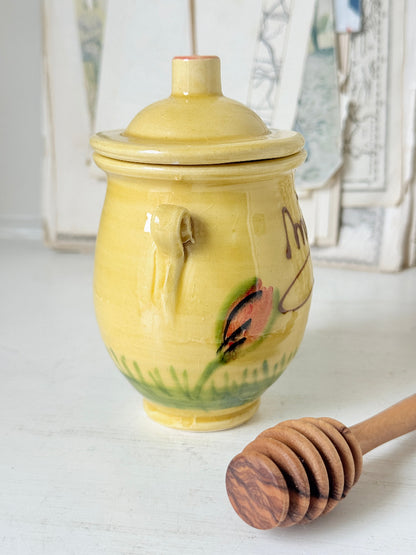 A yellow ceramic honey pot with a lid, featuring a painted bee and green grass design, shown on a white surface with a wooden honey dipper in the foreground.