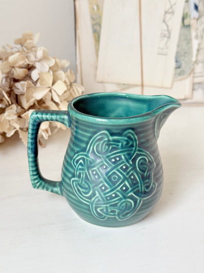 A teal ceramic pitcher with a ribbed handle and a raised Celtic knot pattern on the front, set on a white surface with dried flowers and a blurred background.